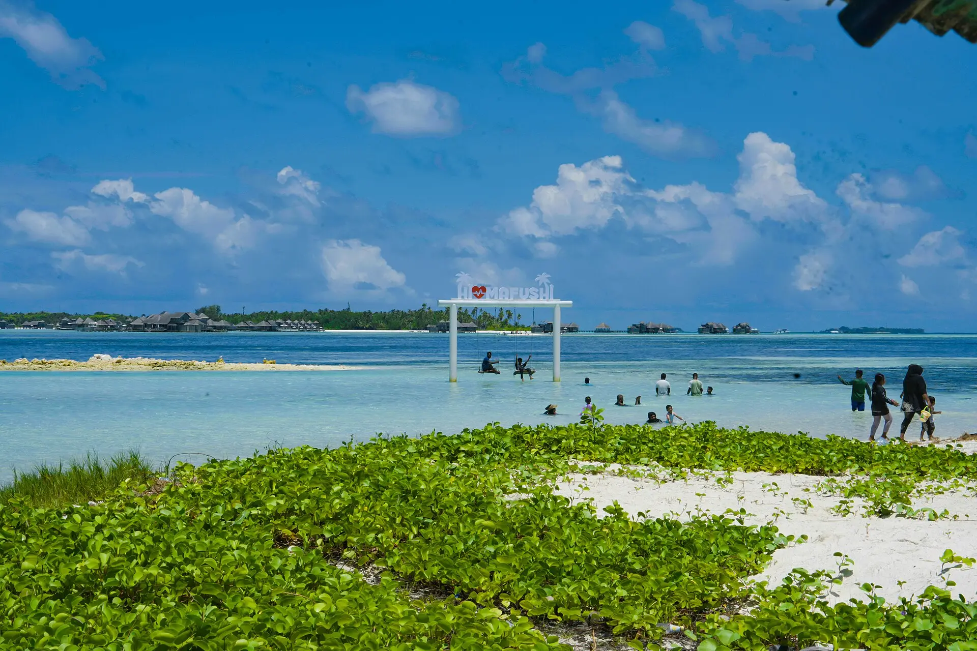 The local beach on Himmafushi with green beach vegetation, a red-and-white swing frame in the shallows and a resort on the horizon