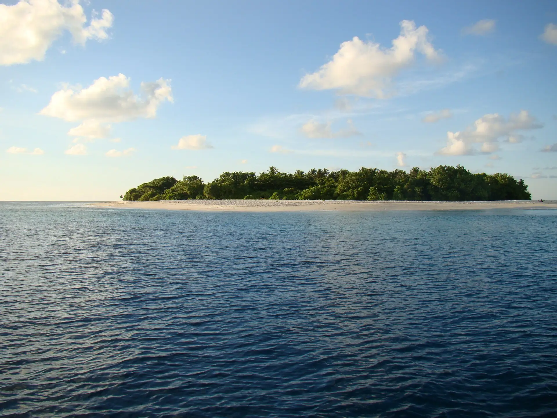 The low green island of Ukulhas with a white sand fringe, seen across deep blue water